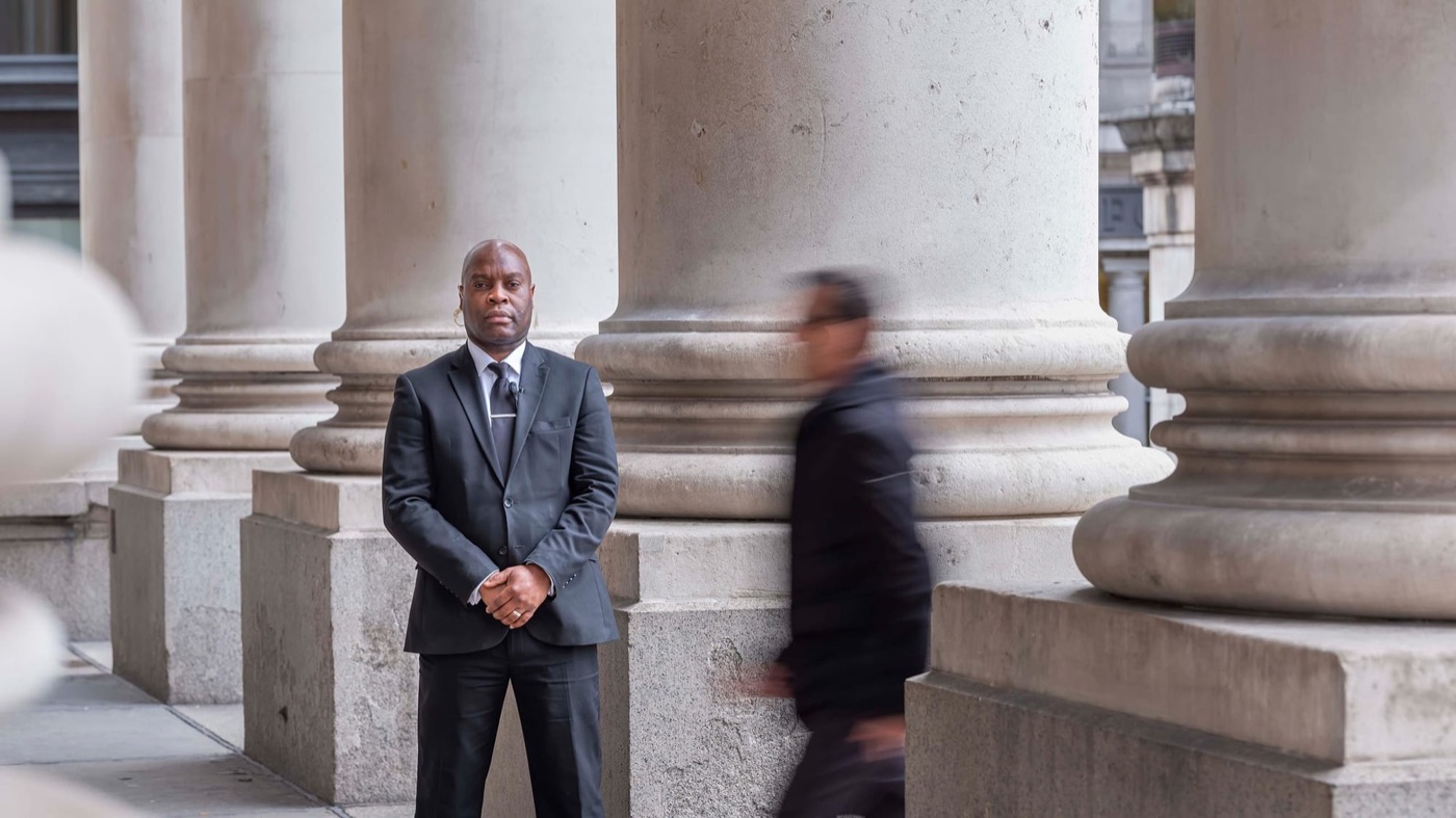 A security officer in a dark suit standing between the stone columns of a city building.