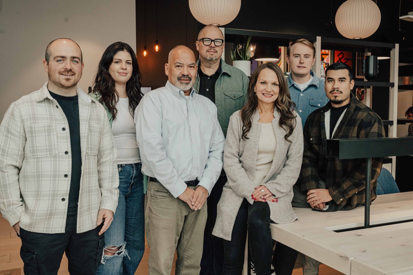 The MTRL team, seven people, standing together in their studio under paper pendant lamps.